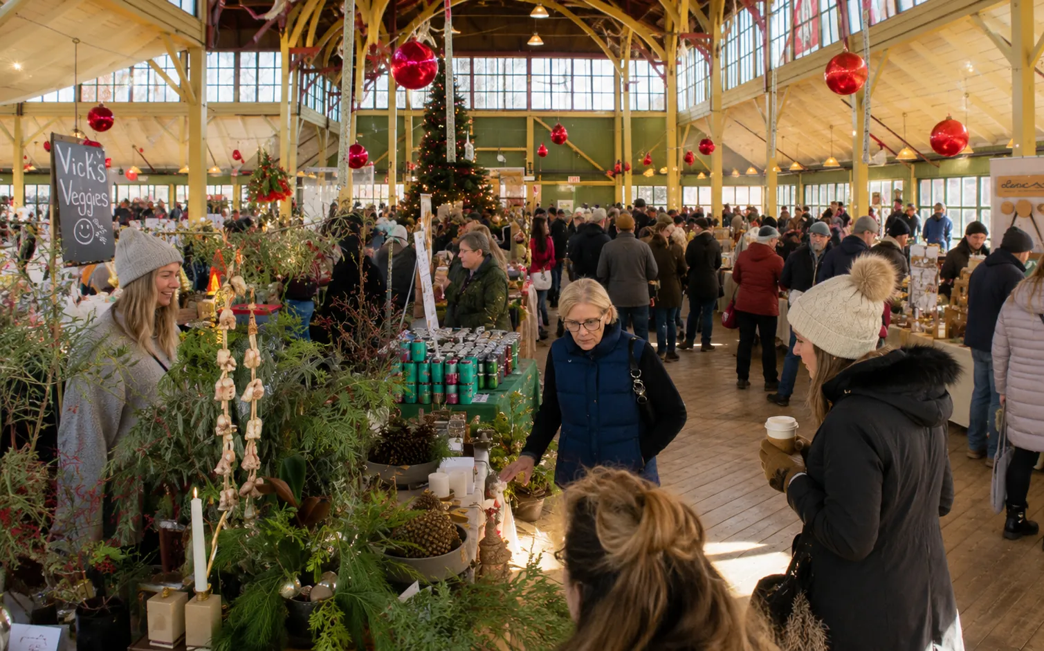 A busy, brightly lit market aisle inside a decorated exhibit hall with shoppers browsing vendor booths surrounded by festive holiday decorations and Christmas trees.